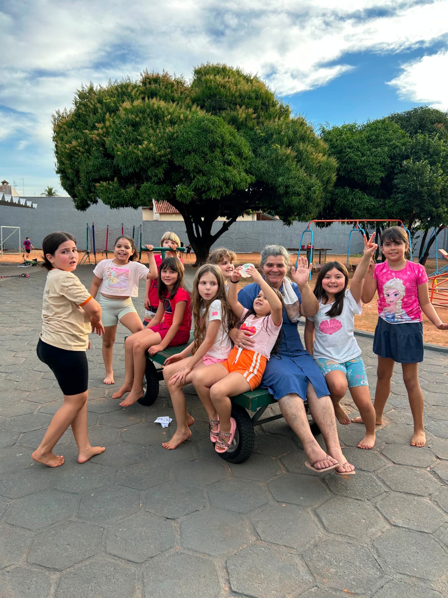 Turma de crianças com uma colaboradora sentadas em um carrinho no pátio, sob árvore frondosa.