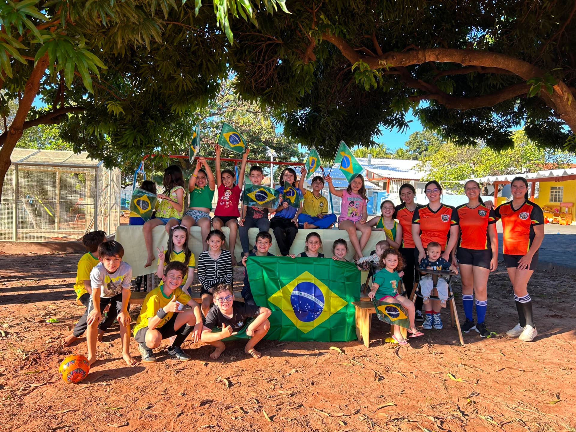 Crianças e equipe reunidas com bandeiras do Brasil em uma programação especial sob a árvore.